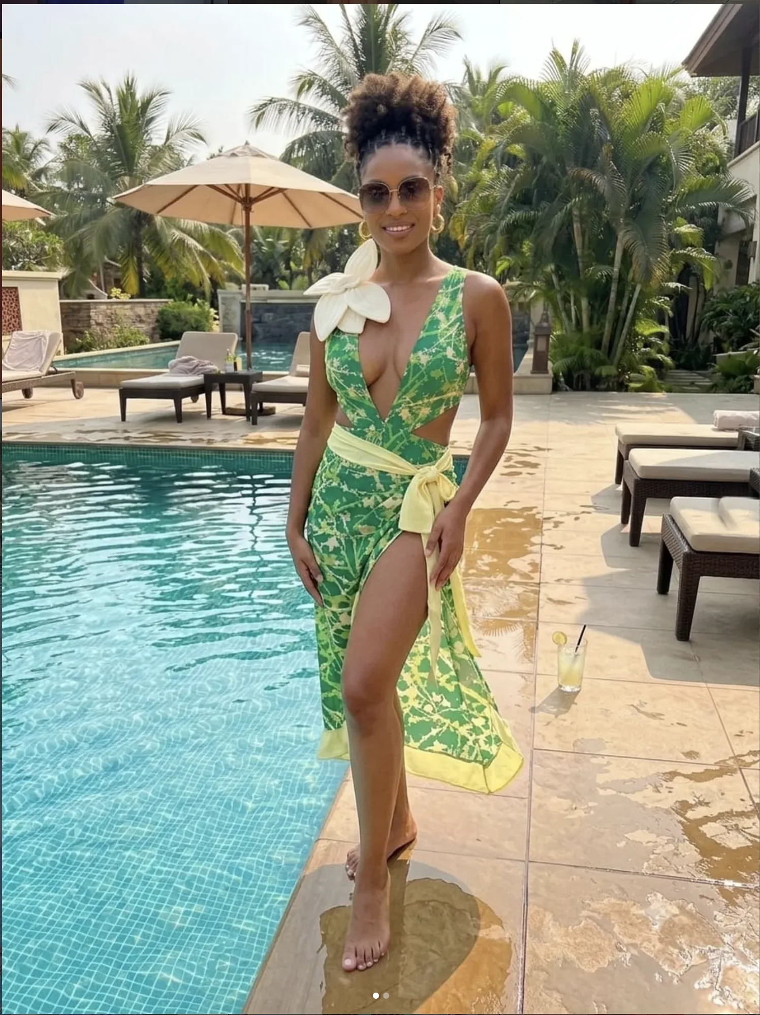 A woman in a green palm-print one-piece and sarong beside a pool framed by palms