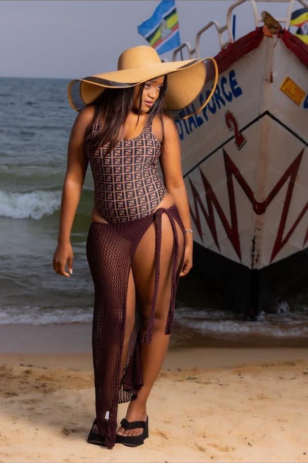A woman in a wide-brimmed straw hat, a monogram-print one-piece and a long burgundy mesh cover-up, walking across the sand beside a painted fishing boat